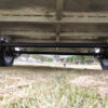 From beneath the Premium 12x6 Dual Axle Heavy Duty Tandem Trailer (3500KG ATM), you can see its metal frame, axles, and wheels. Grass covers the ground with a park-like backdrop of trees.