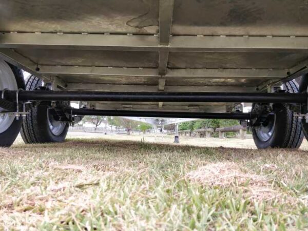 View from beneath the Premium 10x6 Dual Axle Heavy Duty Tandem Trailer - 3200KG ATM For Sale, showcasing its undercarriage, axles, and wheels on grass. The blurred trees and a structure in the background suggest an outdoor setting.
