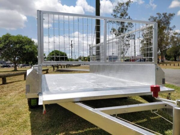 The Premium 12x6 Dual Axle Heavy Duty Tandem Trailer, 3500KG ATM, with a mesh gate and red hitch, sits empty and metallic on grass against a clear blue sky with trees.