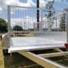 A Premium 10x5 Dual Axle Heavy Duty Tandem Trailer - 3200KG ATM stands in a grassy area with a metal mesh gate. Its made of shiny metal, surrounded by trees, a pole, and under a partly cloudy sky.