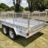 A Premium 8x5 Dual Axle Heavy Duty Tandem Trailer - 3200KG ATM For Sale, featuring galvanized metal mesh sides and white wheels, is parked on grass with trees and a blue sky in the background.