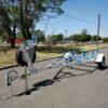 A 4.8m Galvanized Boat Trailer with a 750kg ATM featuring a metal frame and blue rollers sits on a paved road with trees, grass, and benches in the background. A spare tire is attached to the front; power lines and a pole are also visible.