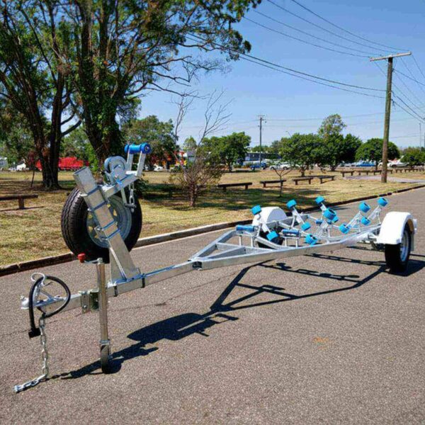 A 4.8m Galvanized Boat Trailer with 750kg ATM, featuring a silver frame, blue rollers, and a mounted spare tire, is parked on a road. Trees, park benches, and electric poles are in the background under a clear sky.