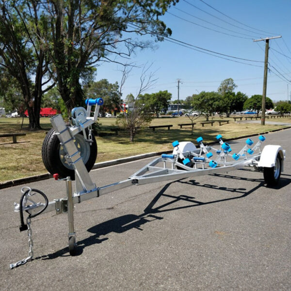 A 4.8m Galvanized Boat Trailer with a 750kg ATM featuring a metal frame and blue rollers sits on a paved road with trees, grass, and benches in the background. A spare tire is attached to the front; power lines and a pole are also visible.
