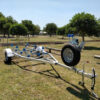 A 4.8m Galvanized Boat Trailer with a 750kg ATM rests on grass in a park. It features a silver frame with blue rollers and a front-mounted spare tire, against a backdrop of trees and a fence under clear skies.