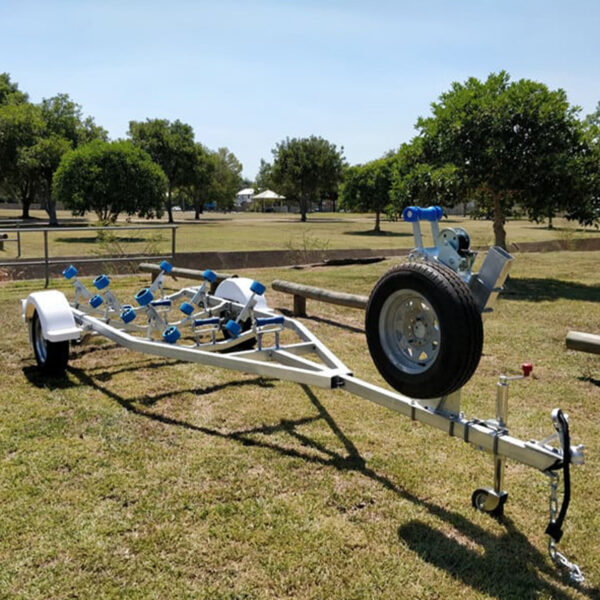A 4.8m Galvanized Boat Trailer with a 750kg ATM rests on grass in a park. It features a silver frame with blue rollers and a front-mounted spare tire, against a backdrop of trees and a fence under clear skies.
