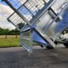 An 8×5 Dual Axle Hydraulic Tipper Tandem Box Trailer - 3500KG ATM With Ramp Holders is tilted on a paved road, against a background of a grassy field and trees. The trailer includes a wire mesh section and diamond plate surface beneath partly cloudy skies.