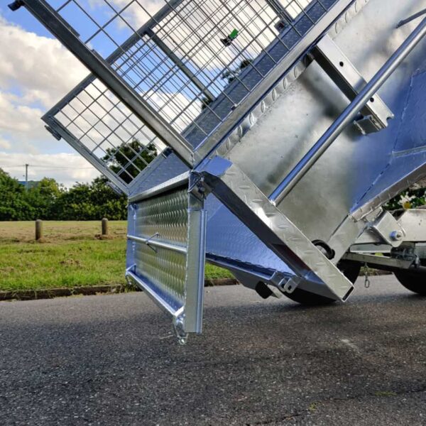An 8×5 Dual Axle Hydraulic Tipper Tandem Box Trailer - 3500KG ATM With Ramp Holders is tilted on a paved road, against a background of a grassy field and trees. The trailer includes a wire mesh section and diamond plate surface beneath partly cloudy skies.