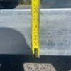 A yellow measuring tape is extended vertically against the metal surface of a 12×6 Dual Axle Livestock/Cattle Trailer with Side Rails and Ramps. It shows measurements in centimeters and inches, casting a shadow on the textured ground. A tire is visible in the upper left corner.