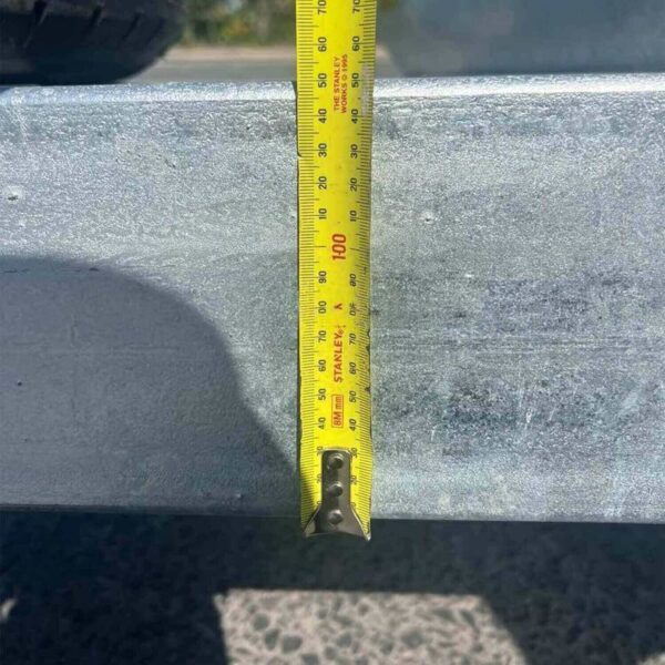 A yellow measuring tape is extended vertically against the metal surface of a 12×6 Dual Axle Livestock/Cattle Trailer with Side Rails and Ramps. It shows measurements in centimeters and inches, casting a shadow on the textured ground. A tire is visible in the upper left corner.