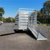 A 12×6 Dual Axle Livestock/Cattle Trailer – 3500KG ATM with side rails and ramps is parked on a paved area, with trees and a vehicle visible in the background under a clear blue sky.