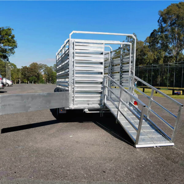 A 12×6 Dual Axle Livestock/Cattle Trailer – 3500KG ATM with side rails and ramps is parked on a paved area, with trees and a vehicle visible in the background under a clear blue sky.