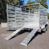 A 12×6 Dual Axle Livestock/Cattle Trailer rated at 3500KG ATM, featuring side rails, open side panels, a roof, and dual ramps for loading on a two-wheel base is parked on a road with trees and greenery in the background.