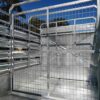 Interior view of a 12×6 Livestock/Cattle Trailer with wire mesh and metal panels. The trailer features a gate with a latch, set against a blue sky and tree branches.