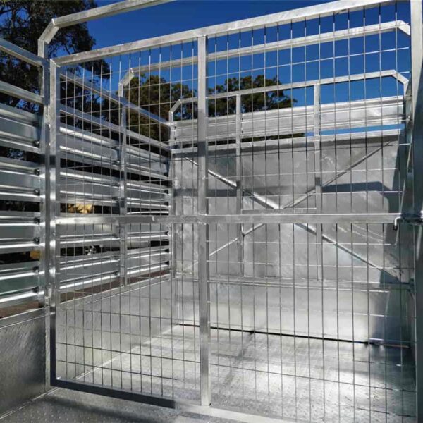 Interior view of a 12×6 Livestock/Cattle Trailer with wire mesh and metal panels. The trailer features a gate with a latch, set against a blue sky and tree branches.