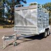 A 12x6 Dual Axle Livestock/Cattle Trailer (3500KG ATM) with side rails and ramps is parked on an asphalt surface, surrounded by trees, under a clear blue sky.