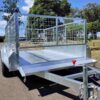A 7×5 Single Axle Box Trailer ATM 1400KG with metal mesh sides and a jockey wheel is parked on a sunny road. It features a shiny metallic finish, a red latch at the front, and is surrounded by trees and grass.