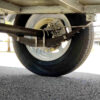 Close-up view of the 7×5 Single Axle Box Trailer ATM 1400KG With Jockey Wheel's suspension system featuring leaf springs, axle, and tire assembly from underneath, with sunlight casting shadows on the asphalt below.
