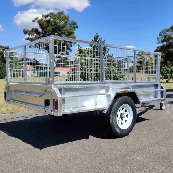 An 8×5 Single Axle Box Trailer ATM 1400KG with Jockey Wheel, metal with high mesh sides, is parked on a paved surface. It has a single axle and white wheel, set against a backdrop of trees, grass, and a partly cloudy sky.