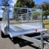 An 8×5 Single Axle Box Trailer ATM 1400KG with a jockey wheel and grid sides is parked on a paved surface, featuring a red handle on the front. Trees and grass are visible in the background under a partly cloudy sky.