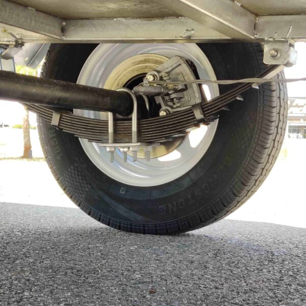Close-up view of the underside of an 8×5 Single Axle Box Trailer ATM 1400KG with Jockey Wheel, showcasing the wheel, suspension, and leaf springs. The trailer sits on asphalt under bright sunlight, casting soft shadows.