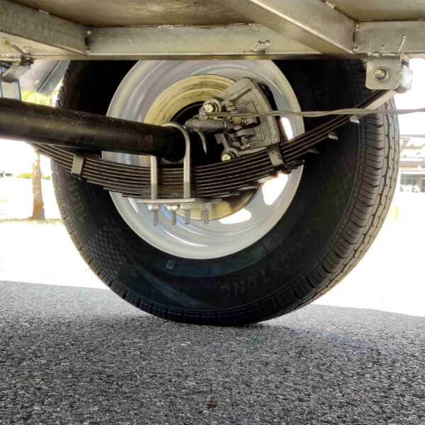 Close-up of the rear wheel and suspension system on the 8×5 Single Axle Box Trailer ATM 1400KG With Jockey Wheel, featuring a leaf spring, axle, and tire, with sunlight in the background.