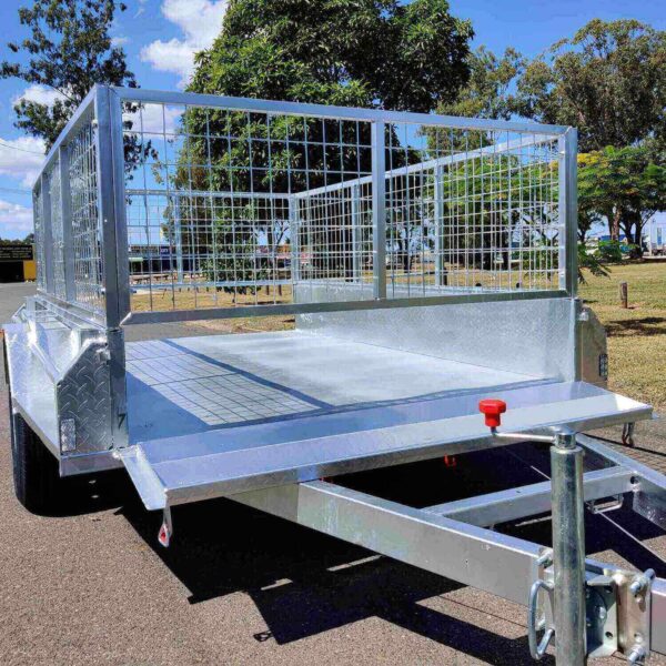 A 9×5 Single Axle Box Trailer ATM 1400KG with a jockey wheel is parked on a paved road. It has high mesh sides, dual wheels, and a flatbed design ideal for transporting goods. Trees and a partially cloudy sky are in the background.