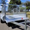 A 9×5 Single Axle Box Trailer ATM 1400KG with a jockey wheel and mesh sides is parked on a paved road. It features a red handle at the front for hitching, surrounded by trees and grass under a partly cloudy sky.
