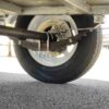A close-up view shows the 9×5 Single Axle Box Trailer ATM 1400KG with a jockey wheel from underneath, highlighting its suspension system with leaf springs, axle, and brake caliper. The vehicle rests on pavement with trees visible in the background.