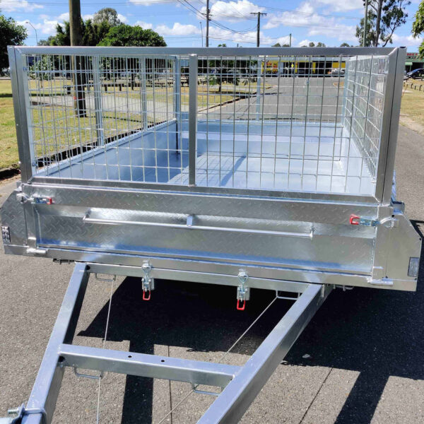 A 9×5 Single Axle Box Trailer ATM 1400KG With Jockey Wheel, featuring a galvanized steel frame, mesh sides, diamond plate floor, and rear latch mechanisms, is empty and parked on a paved road with trees and other trailers in the background.