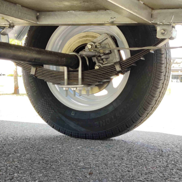 Close-up view under the 9×5 Single Axle Box Trailer ATM 1400KG With Jockey Wheel, showing its tire, metal rim, leaf spring suspension, and axle components on a paved surface.