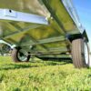 A close-up of the 7×4 Single Axle Box Trailer ATM 750KG with Jockey Wheel showcases its metallic underside and two large black tires on green grass, set against a clear blue sky in the background.