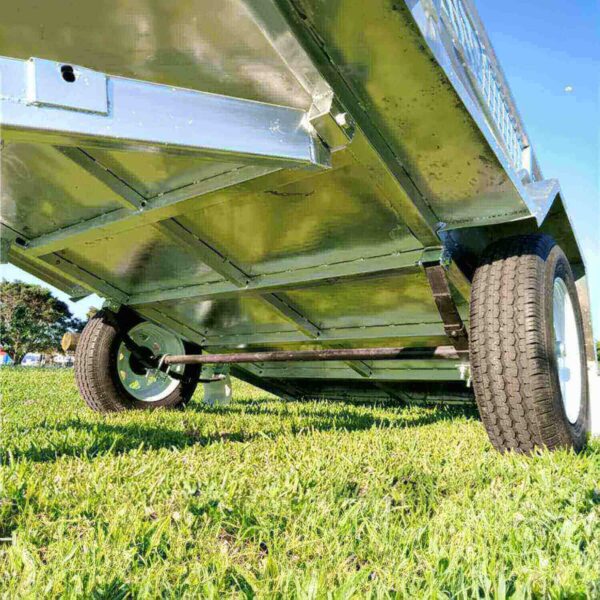 A close-up of the 7×4 Single Axle Box Trailer ATM 750KG with Jockey Wheel showcases its metallic underside and two large black tires on green grass, set against a clear blue sky in the background.
