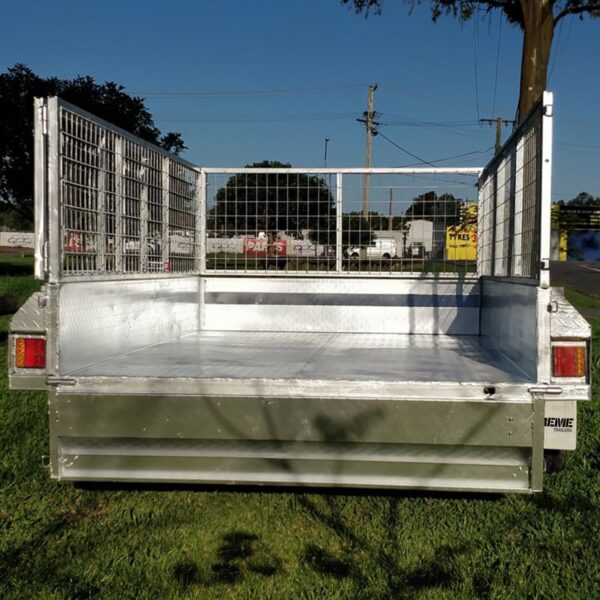 A 7x4 Single Axle Box Trailer ATM 750KG with a jockey wheel and metal mesh sides is parked on grass. It is empty, with a road, power lines, and trees visible in the background under a clear blue sky.
