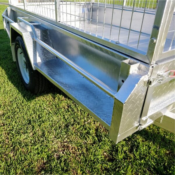 The 7×4 Single Axle Box Trailer ATM 750KG with a wire mesh top and diamond plate flooring stands on grassy terrain, its shiny metallic surface highlighted by sunlight, with one wheel visible.