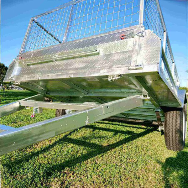 Low-angle view of a metallic 7×4 Single Axle Box Trailer ATM 750KG With Jockey Wheel on grass, showcasing a diamond plate surface with mesh upper, red latch detail, black wheels, and an extended hitch in the foreground.