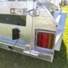 Close-up of a 7×4 Single Axle Box Trailers rear corner, featuring a red and orange taillight, metal pin latch, and a black box with wire. Parked on grass, sunlight reflects off its metallic surface.