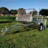 A 7×4 Single Axle Box Trailer ATM 750KG With Jockey Wheel, featuring a silver cage, two wheels, protective mesh, and a hitch for towing, sits on a grassy field. Trees and buildings are visible in the background under the clear blue sky.