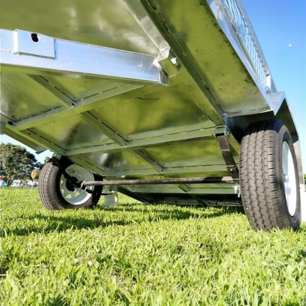 Close-up view of a 7x5 Single Axle Box Trailer ATM 750KG with Jockey Wheel, set on green grass, showcasing the underside with visible axle and two tires; sunlight creates shadows beneath while trees and sky appear blurred in the background.
