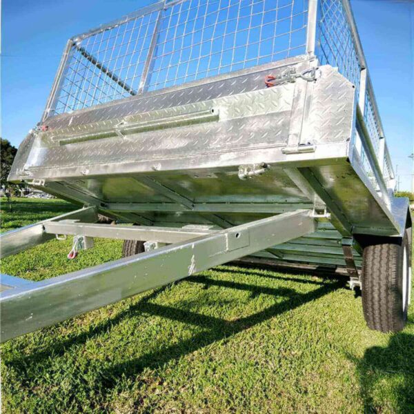 Close-up of the 7x5 Single Axle Box Trailer ATM 750KG With Jockey Wheel on grass, showcasing its metallic frame, mesh gate, and two wheels. The image highlights the sturdy construction and diamond plate texture under a sunny, clear sky.