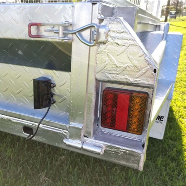 Close-up of a metallic corner of the 7x5 Single Axle Box Trailer ATM 750KG with Jockey Wheel on grass, showing a diamond plate pattern, red and orange tail light, small black wired device, and latch with looped pin for securing the side panel.