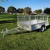 A 7x5 Single Axle Box Trailer ATM 750KG with a jockey wheel and mesh sides sits on a grassy field. It features a single axle with a black tire and tow hitch at the front. Trees and a distant road are visible in the background under a clear sky.