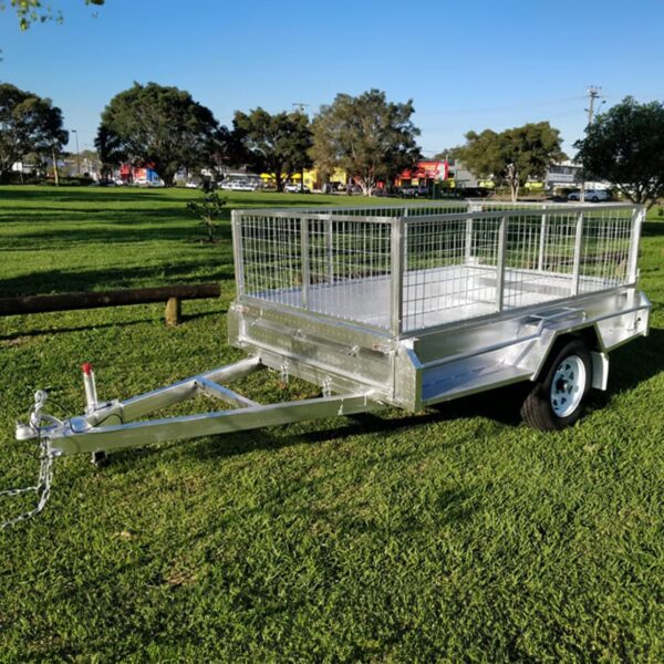 A 7x5 Single Axle Box Trailer ATM 750KG with a jockey wheel and mesh sides sits on a grassy field. It features a single axle with a black tire and tow hitch at the front. Trees and a distant road are visible in the background under a clear sky.
