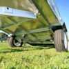 Close-up shot of the underside of an 8x5 Single Axle Box Trailer ATM 750KG With Jockey Wheel in a grassy field, showcasing visible axles and wheels. Sunlight highlights its metallic surfaces, with trees and a blue sky in the background.