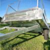 A low-angle view of a 6x4 Single Axle Box Trailer ATM 750KG with a gridded tailgate and Jockey Wheel, parked on green grass. The shiny metal trailer has two wheels and a sturdy frame, reflecting sunlight under a clear blue sky.