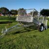 An 8x5 Single Axle Box Trailer ATM 750KG with a mesh cage, tow bar, and jockey wheel is parked on grass under a clear sky. Black tires are on its single axle, and trees plus distant structures can be seen in the background.