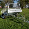 A 6x4 Single Axle Box Trailer with a silver finish, cage, two wheels, and a jockey wheel is parked on green grass. A tow bar extends toward the viewer, with trees and a cloudy blue sky in the background.