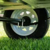 A close-up of the 6x4 Single Axle Box Trailer ATM 750KG with Jockey Wheel shows a black tire with a white rim, secured by a metal U-bolt clamp against green grass. The metal components of the trailers underside are visible in the background.