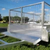 A 6x4 Single Axle Box Trailer ATM 750KG with Jockey Wheel, featuring a mesh wire enclosure, is parked on grass with its back gate open. The sky is mostly clear with some clouds, and trees are seen in the background.