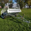 A 6x4 Single Axle Box Trailer ATM 750KG with Jockey Wheel, featuring wire mesh sides, is parked on grass in a park. It has two wheels and a long tow bar with a hitch at the front. Trees and a building are in the background under partly cloudy skies.